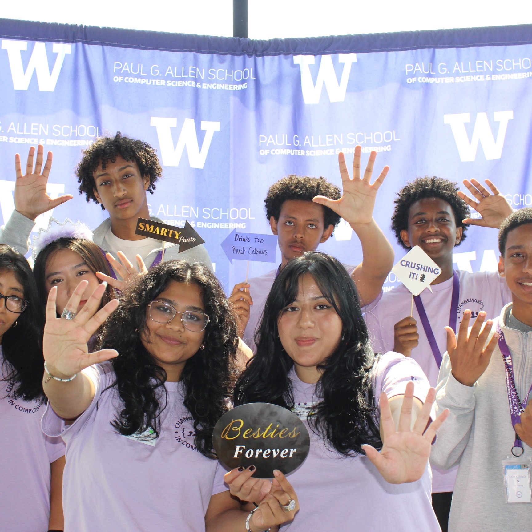 Six highschool students and two young adults pose in a photobooth in front of a purple "Paul G. Allen School" backdrop. They are all smiling and wearing matching lavender t-shirts while folding up five fingers. Some of them are holding up photobooth props with funny phrases ('Besties Forever', 'Smartie Pants', 'Crushing it!', 'Drinks too much Celsius').