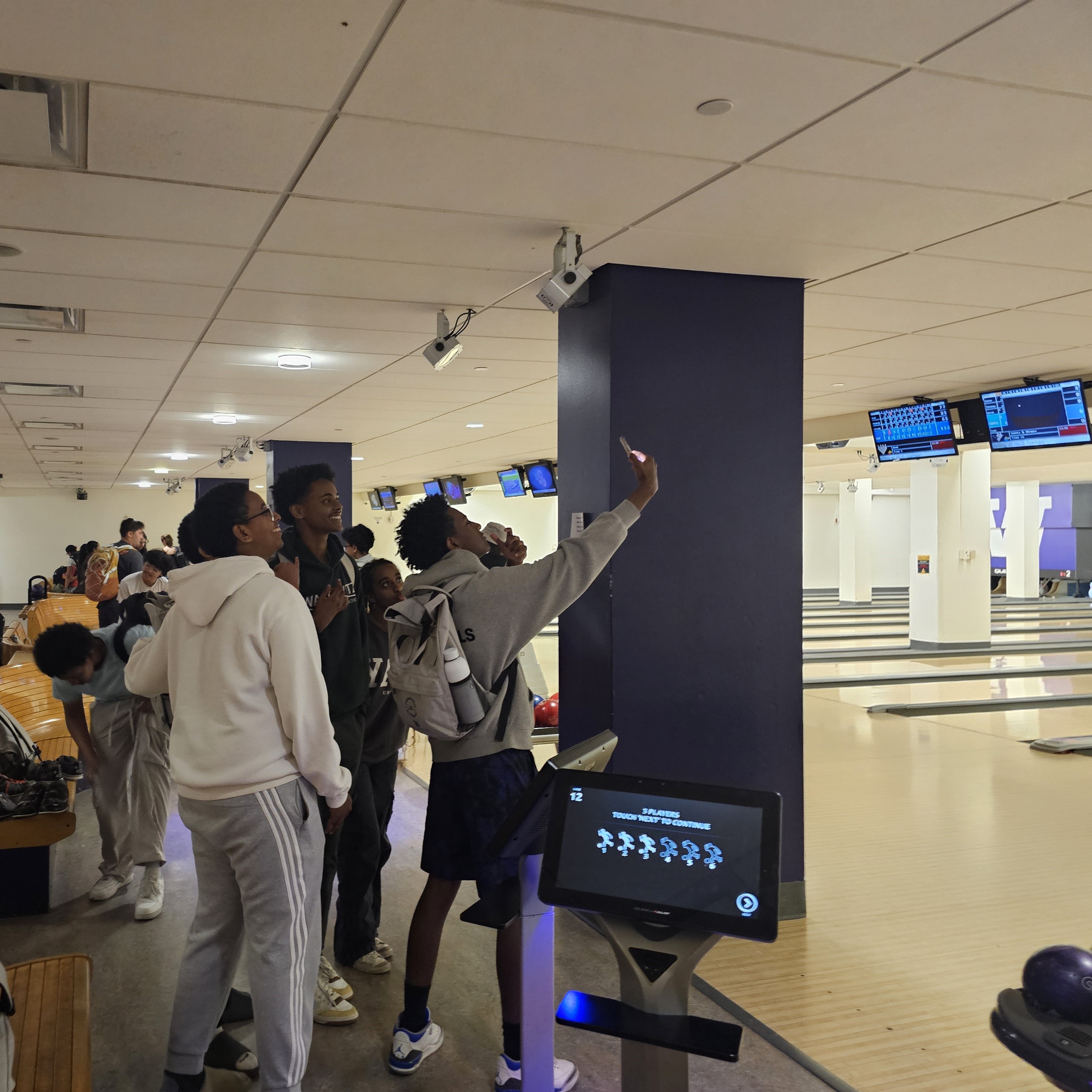 A group of 4 highschool students candidly pose for a phone selfie. They are facing away from the camera and smiling up at their own phone. Behind them is a bowling alley with many lanes.