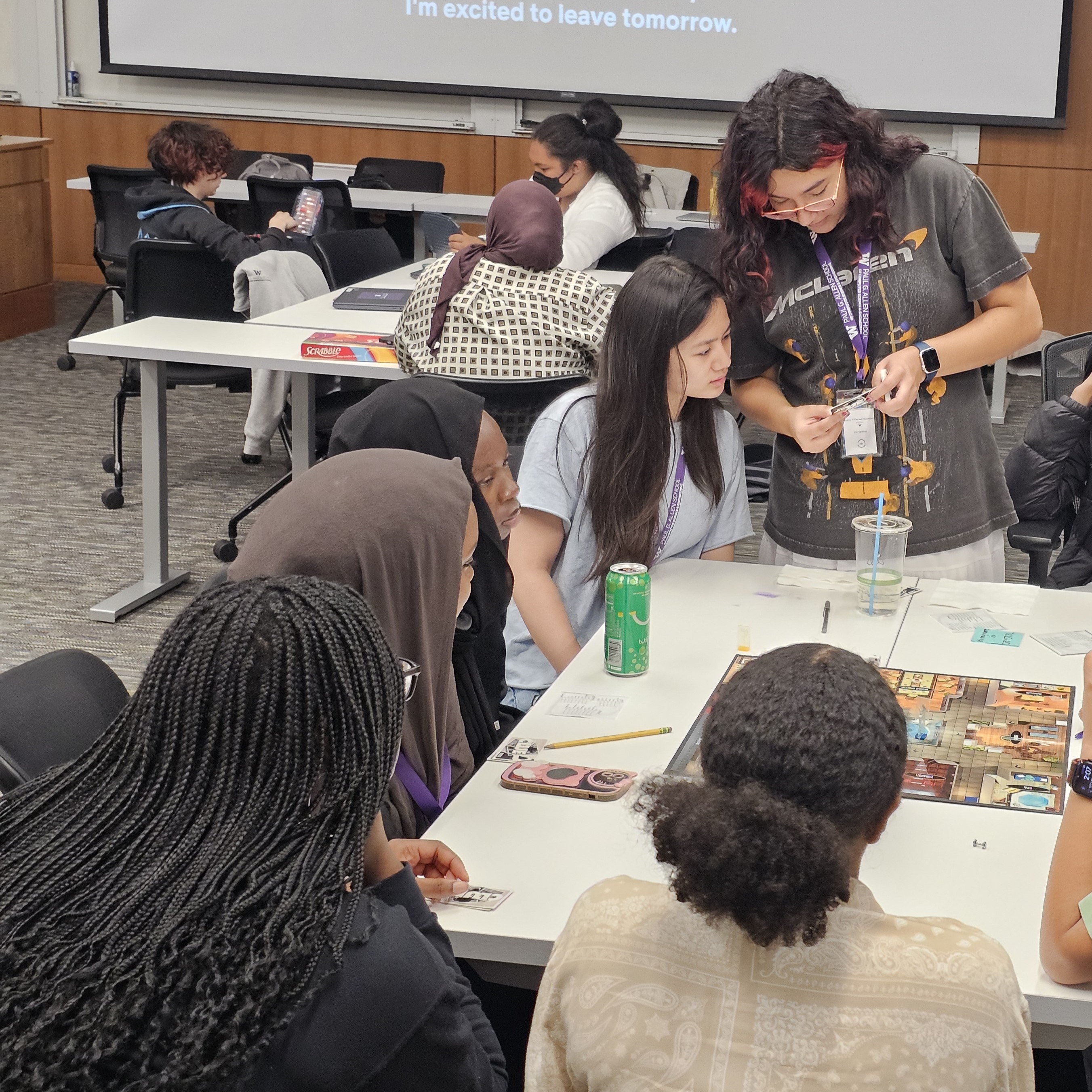 : A group of teenage girls huddle around a large white table with the board game Clue laid on it. They are looking away from the camera, each holding colorful game cards and talking to each other.