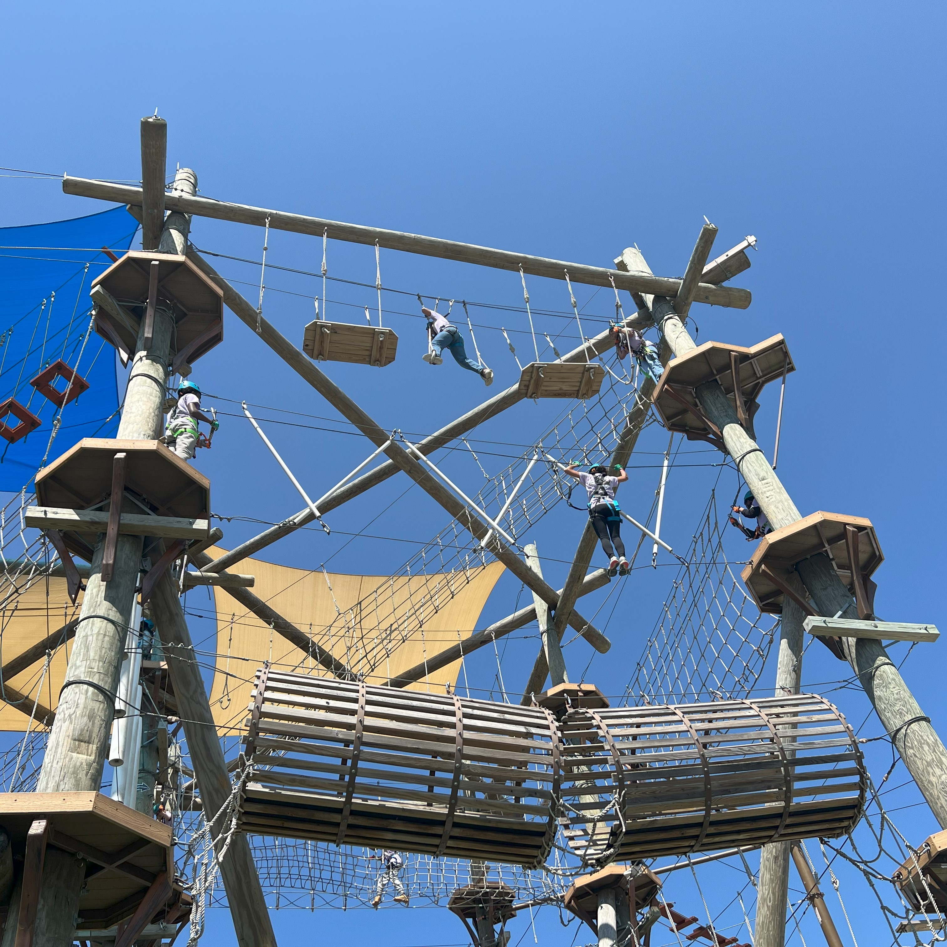  Several students are climbing on a tall ropes course. The students are wearing matching lavender shirts, blue helments, and black harnesses. The ropes course is a wooden structure with three tiers. There are ropes, nets, platforms, ziplines, and other obstacle elements on the course. In the background, the sky is clear and blue.