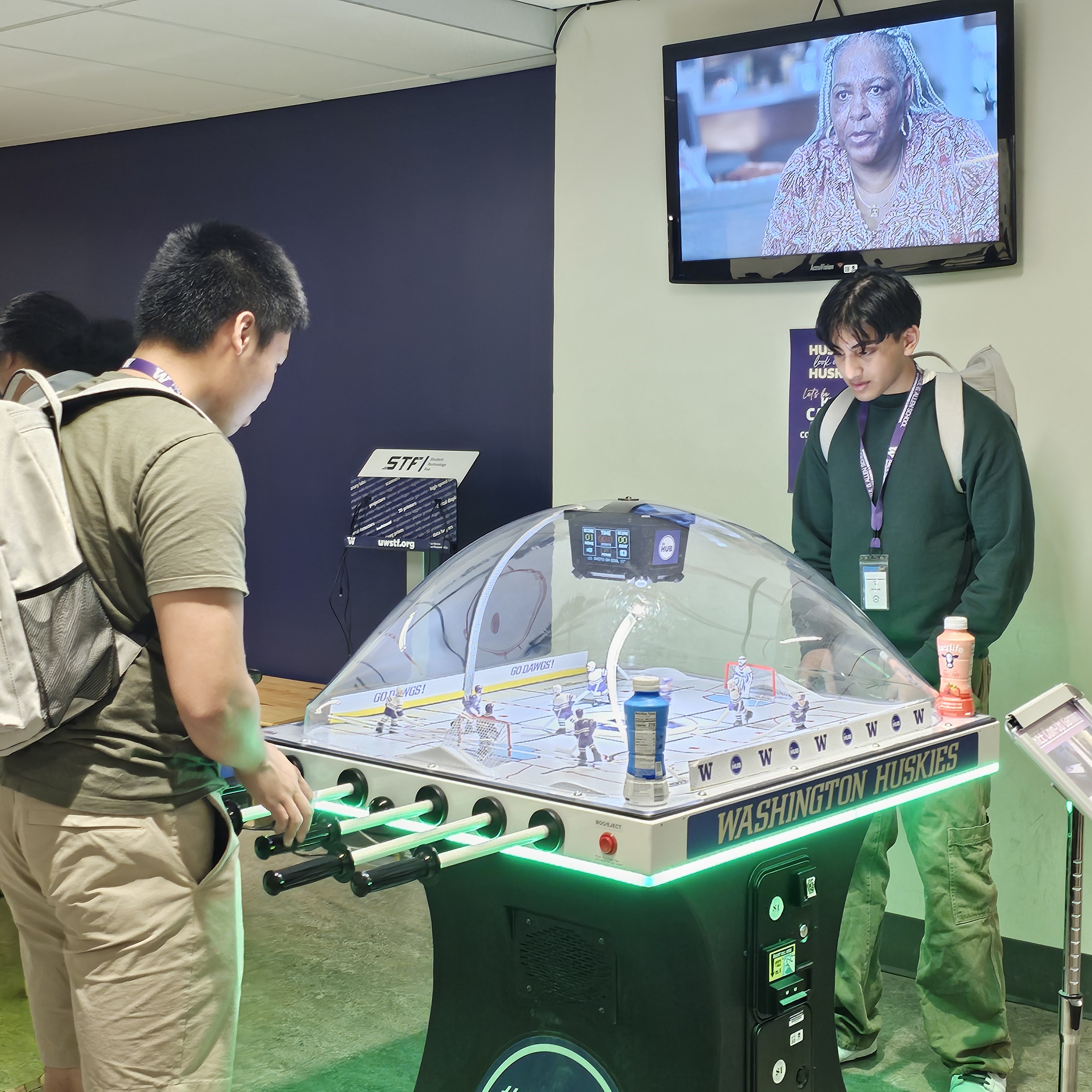 Two highschool students are in an arcade, bent over a table hockey game set with a domed lid. They are facing away from the camera and are looking down towards the game. The set is branded with a University of Washington Huskies logo and the table surrounded by a green LED light.