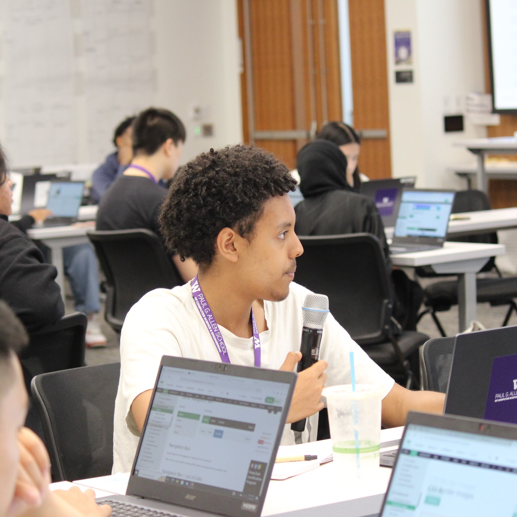 A male highschool student in a lecture hall holding a microphone and answering a question, looking off into the distance.  The student is wearing a white shirt and a purple lanyard, he has brown skin and short dark coily hair. Other students are working on their laptops in the background, but the background is hazy.