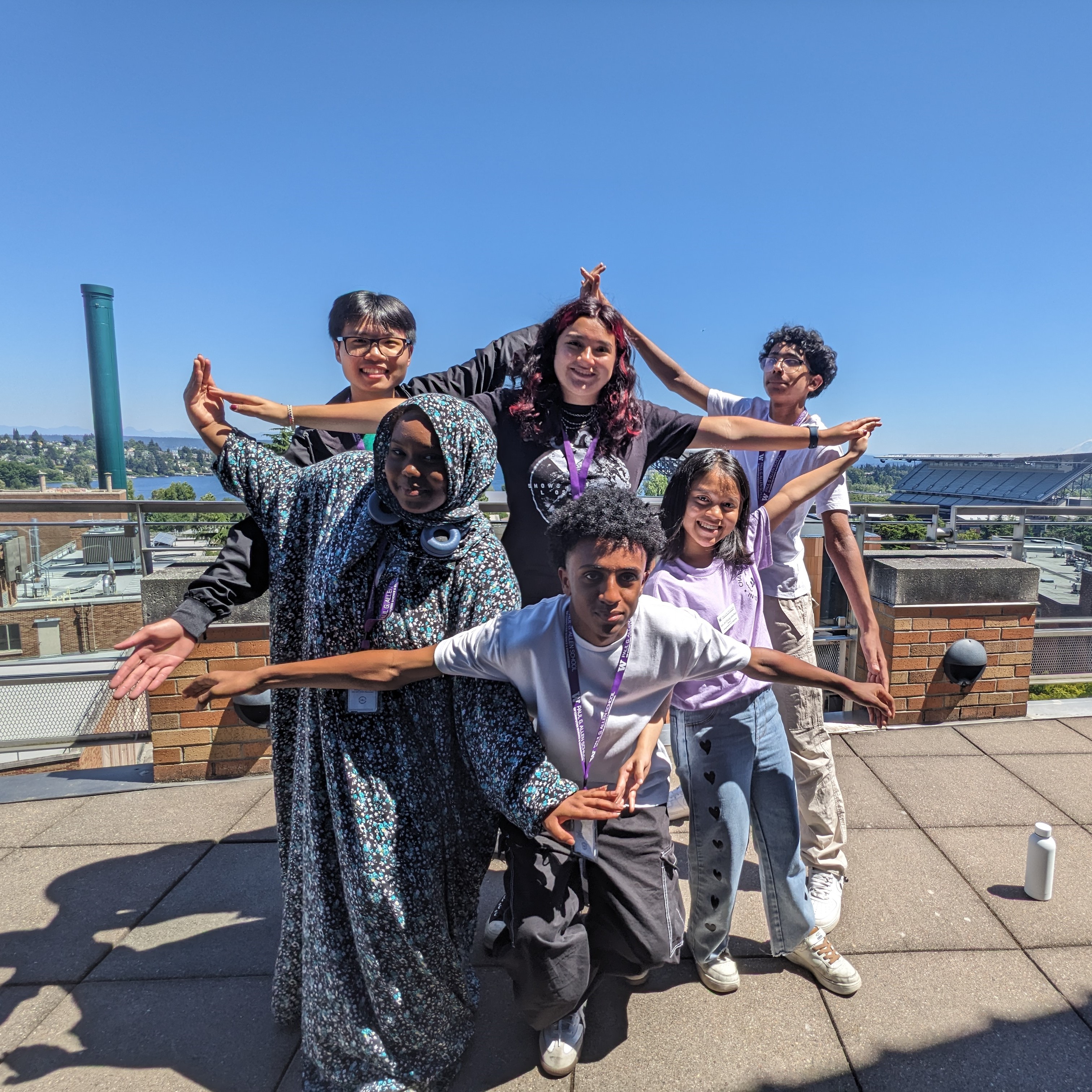 Six highschool students pose on a balcony for a photo with their arms outstretched and pointing together in the shape of a star. They are smiling. Behind them is a clear blue sky and a view of the University of Washington Husky Stadium.