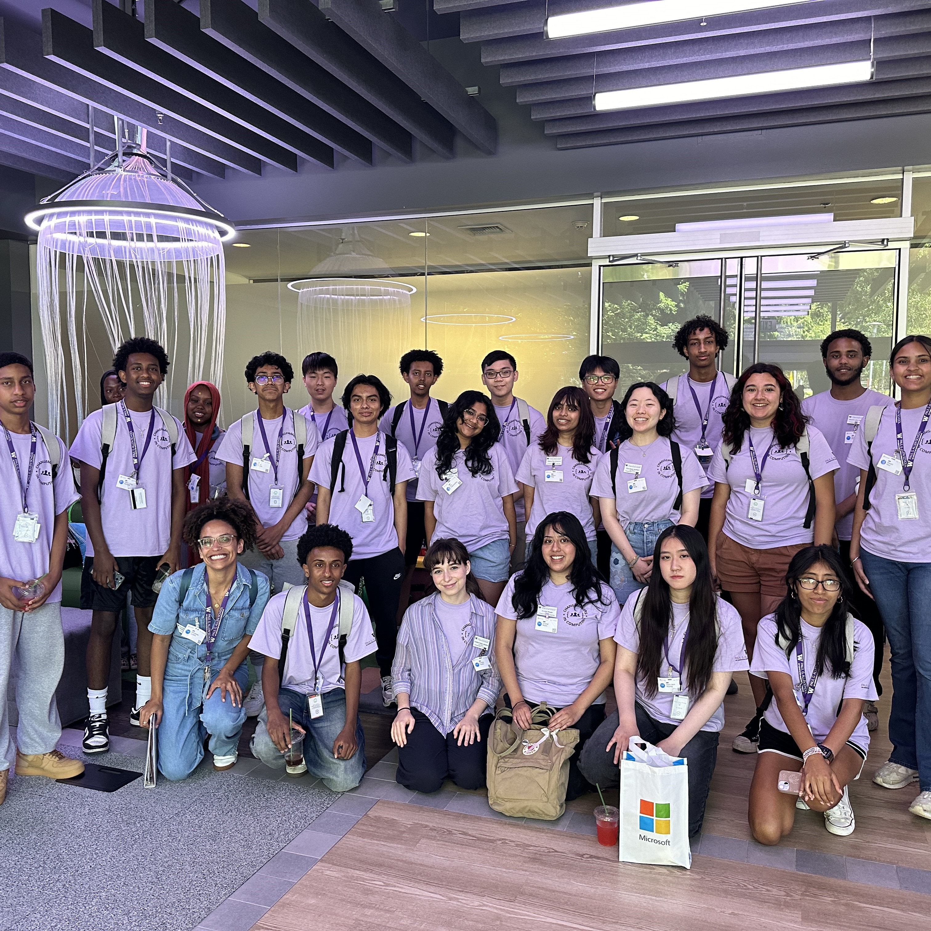 About 20 students pose for a group photo. They are smiling and wearing matching lavender t-shirts and purple lanyards. Behind them is a lab with glass doors and walls and a hanging light fixture that resembles a jelly fish.