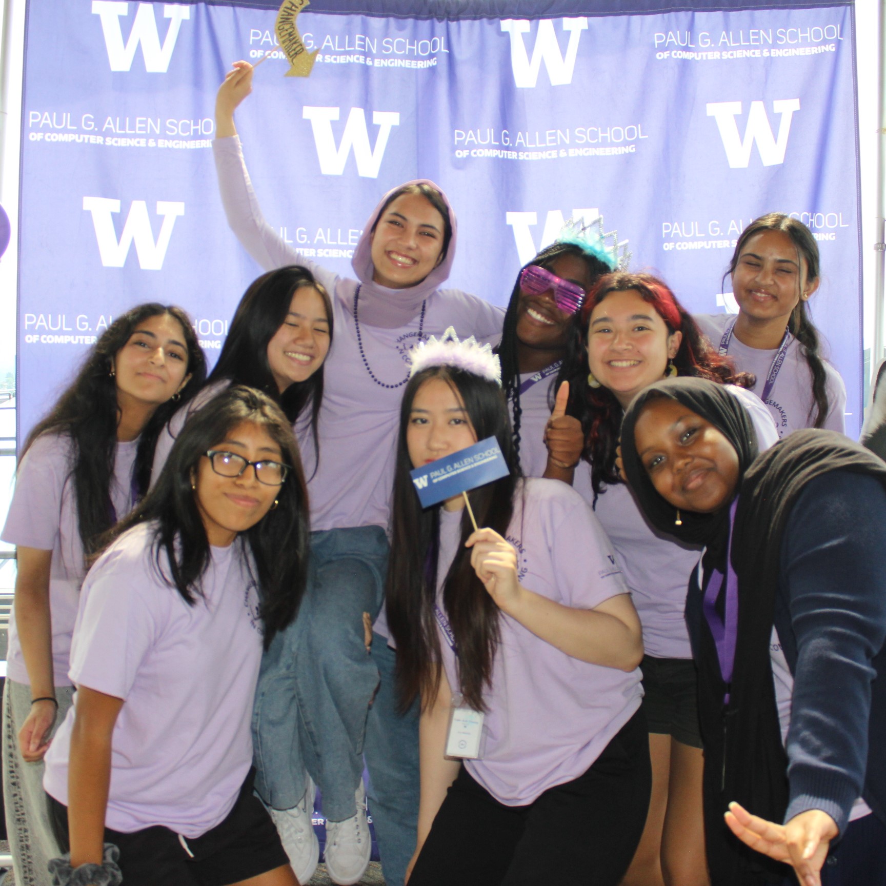 A group of 9 female highschool students strike a fun, joyful pose in a photobooth in front of a purple "Paul G. Allen School" backdrop. They are all smiling and laughing while wearing matching lavender t-shirts. Some of them are wearing festive photobooth props like tiaras, sunglasses, and necklaces. They are holding up thumbs-ups and peace signs as well a sign that says 'Changemaker.'