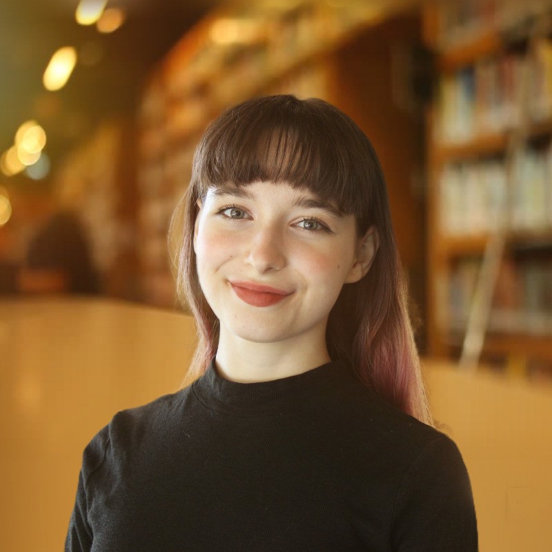 A headshot of Andrea, a young white woman with dark brown hair. She wears a black turtleneck sweater and is smiling at the camera. There is a library in the background.