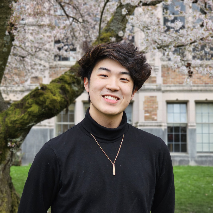 A headshot of Colin. Colin is a 19-year-old Asian American man with black hair. He smiles at the camera wearing a black turtleneck with a gold necklace. There are cherry blossom trees in the background.