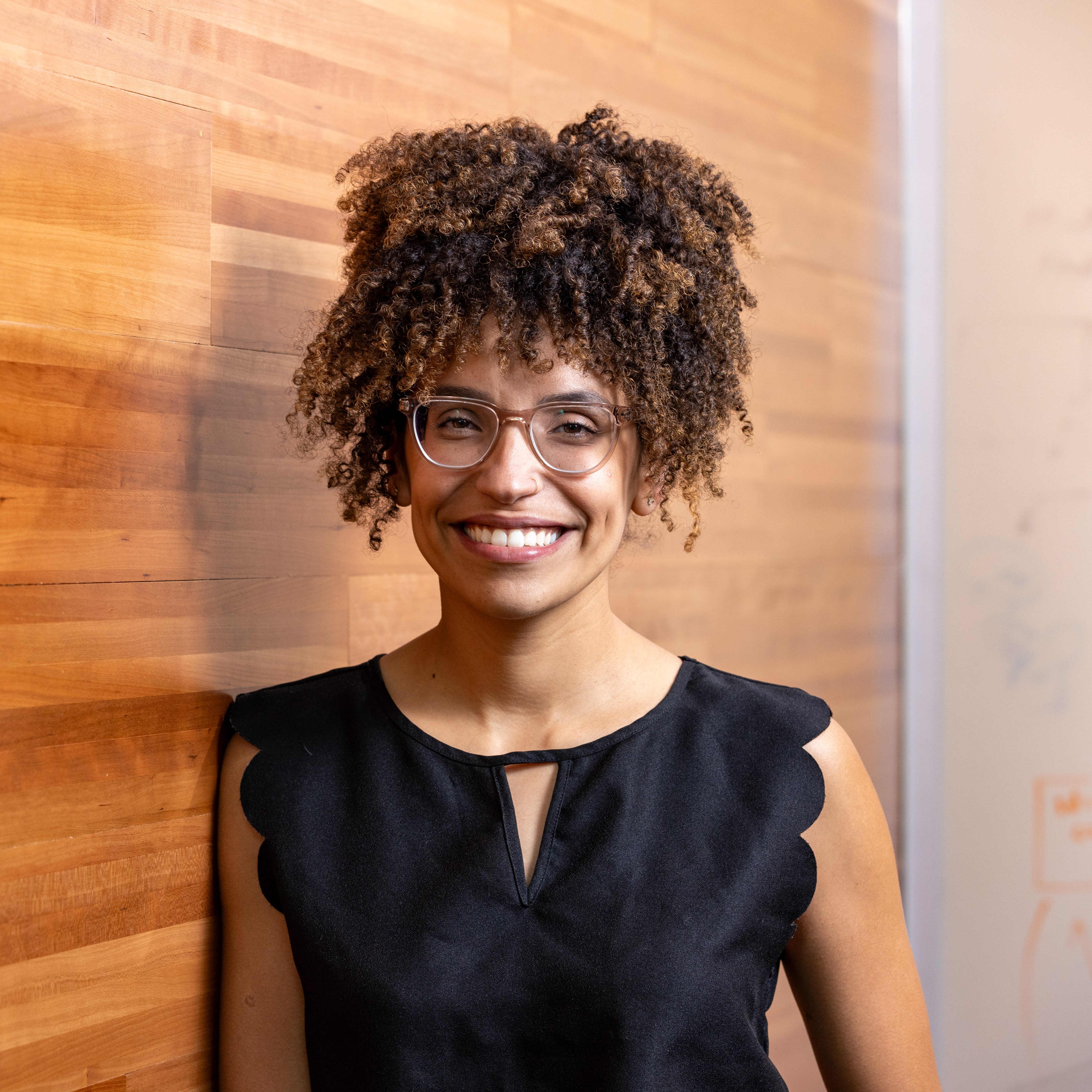 Fernanda is a light-skinned Black Latina woman, looking at the camera and smiling. She wears glasses and has big curly hair. She is wearing a shitless black top and is leaning on a wooden wall.