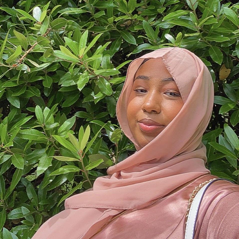 a headshot of Siham, a 20 year old black woman wearing a pink headscarf and a pink dress.